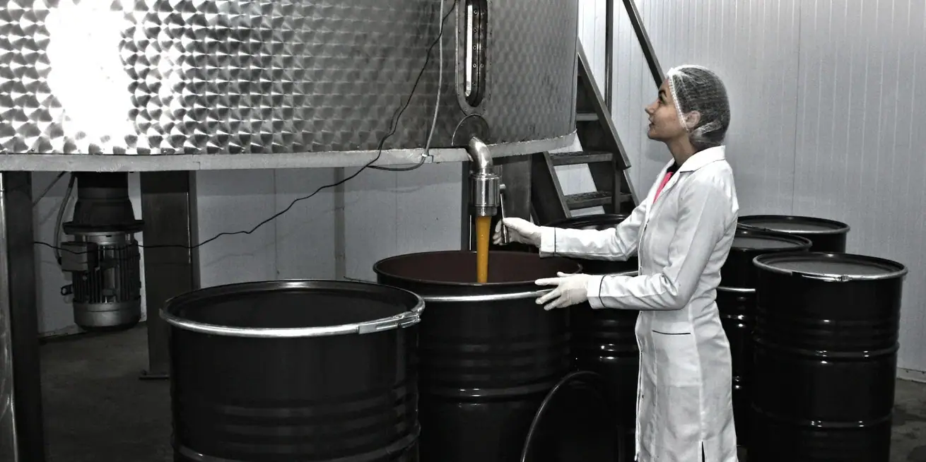 A technician in a lab coat and hairnet filling a steel drum with honey from the homogenising tank.
